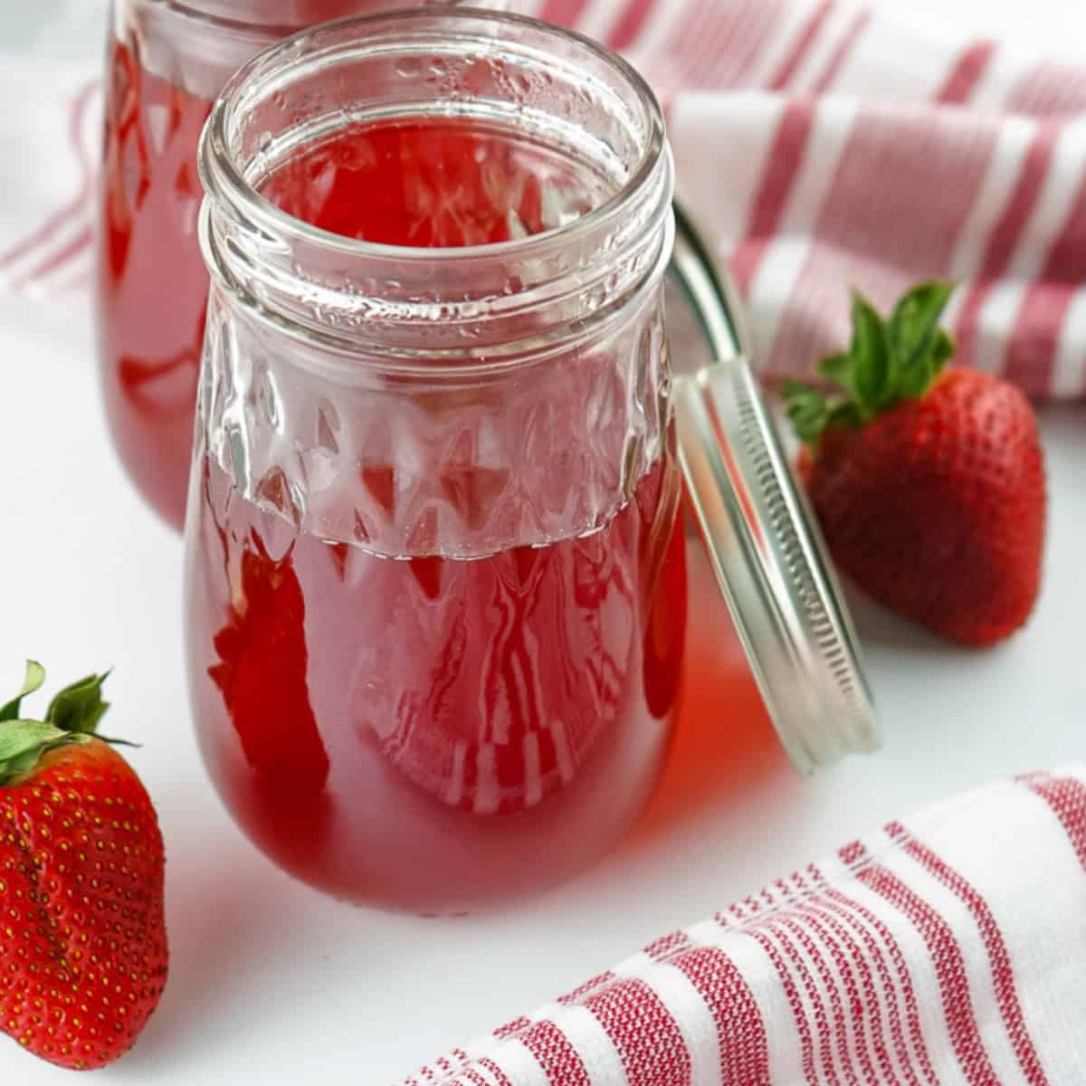Strawberry simple syrup used for making beverages and topping ice cream in a glass jar on a white table with red and white striped linens and fresh strawberries